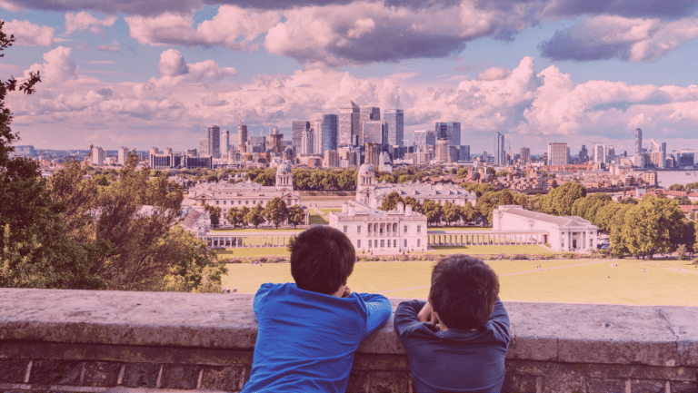 Dois meninos estão apoiados em um muro com vista para o parque Greenwich, em Londres, na Inglaterra. O céu está cheio de nuvens brancas e é possível ver o rio Tâmisa e os prédios de Canary Wharf no horizonte.