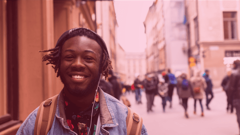 A imagem mostra um homem sorrindo para a câmera enquanto ouve música ao ar livre em um ambiente urbano. Ele está vestindo roupas da moda de rua.