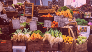 Foto do Mercado Rainha Vitória, na Austrália, mostra diferentes legumes e verduras coloridos e os seus respectivos preços em cestos marrons. Foto de Somi Jaiswal (@somijaiswal_fotografia)